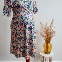 Floral maternity dress on a mannequin against a wall with a vase of dried flowers on a small table to the side