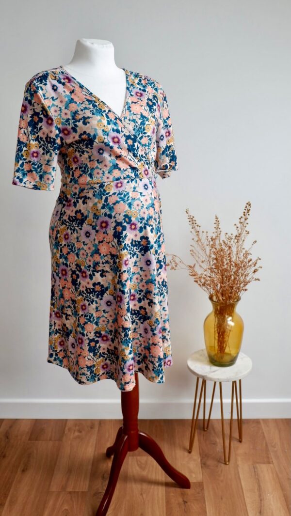 Floral maternity dress on a mannequin against a wall with a vase of dried flowers on a small table to the side