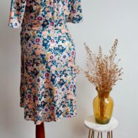 Floral maternity dress on a mannequin showing the back of the dress against a wall with a vase of dried flowers on a small table to the side