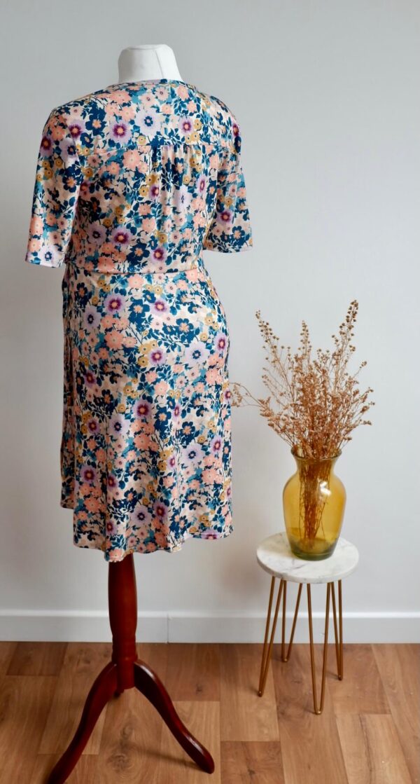 Floral maternity dress on a mannequin showing the back of the dress against a wall with a vase of dried flowers on a small table to the side