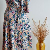 Floral maternity dress on a mannequin showing the side of the dress against a wall with a vase of dried flowers on a small table to the side