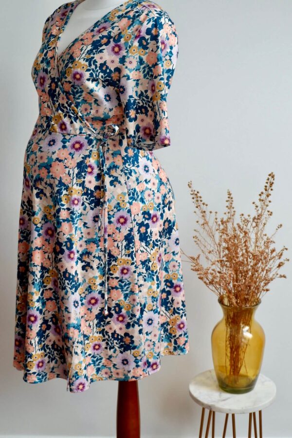 Floral maternity dress on a mannequin showing the side of the dress against a wall with a vase of dried flowers on a small table to the side