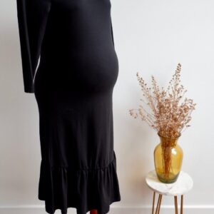 Black dress on a pregnant mannequin in front of a white wall with a small round table to the side with dried flowers in it