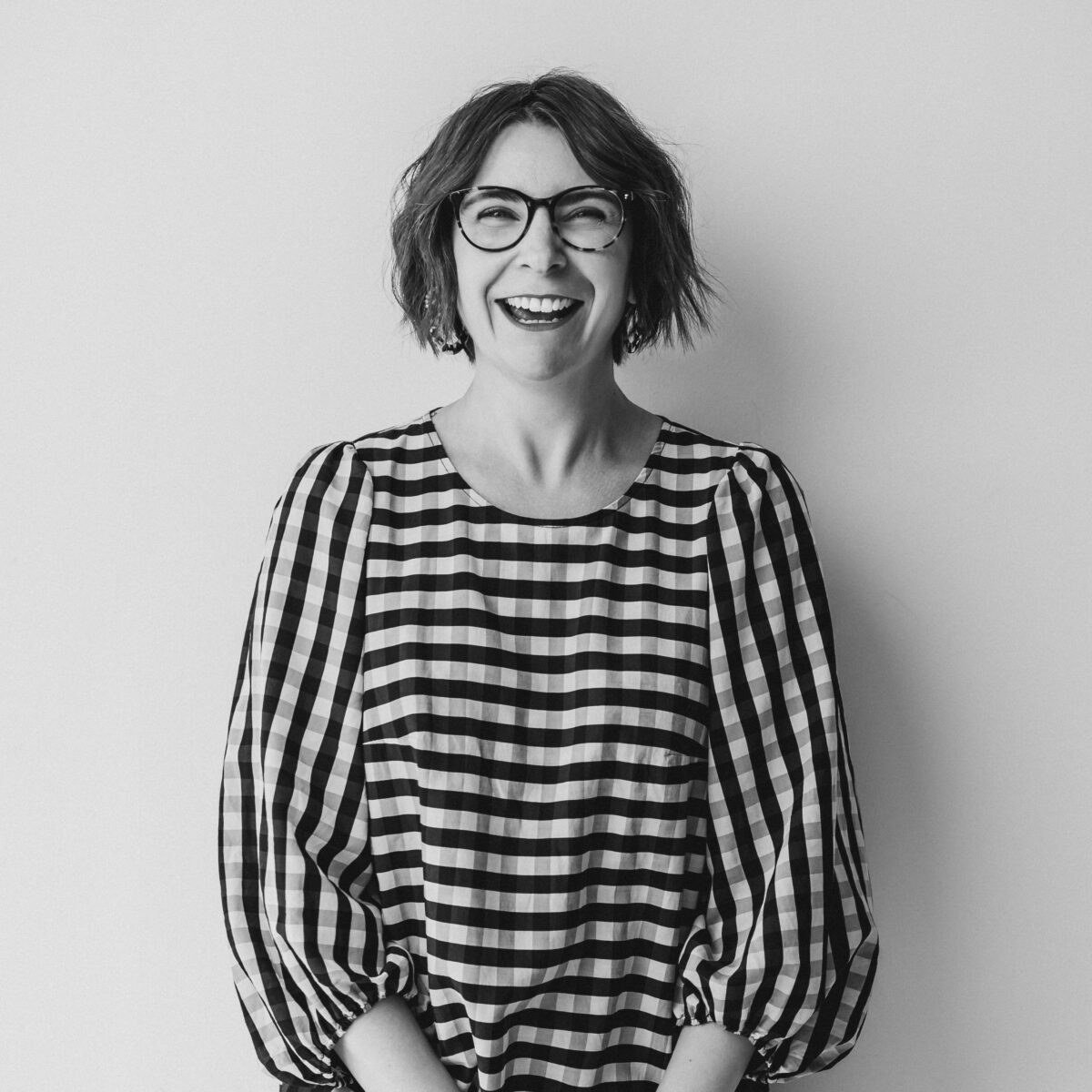 Black and white image of Jo Leggiero, founder of Mother Dragon in a check straight dress with balloon sleeves leaning again a wall looking directly at the camera laughing.