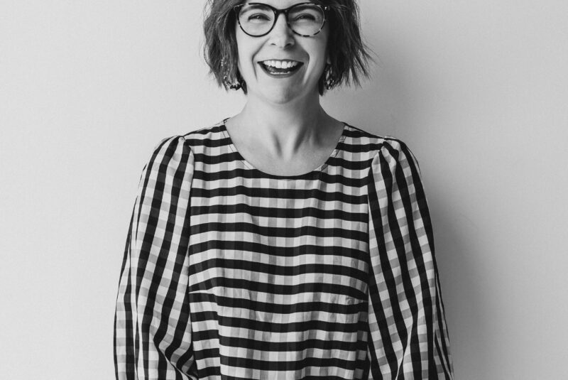 Black and white image of Jo Leggiero, founder of Mother Dragon in a check straight dress with balloon sleeves leaning again a wall looking directly at the camera laughing.
