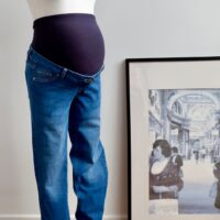 Pregnant mannequin with blue over the bump maternity jeans to the left of the imagine against a white wall. Framed picture to the right of the mannequin rests on the floor against the wall