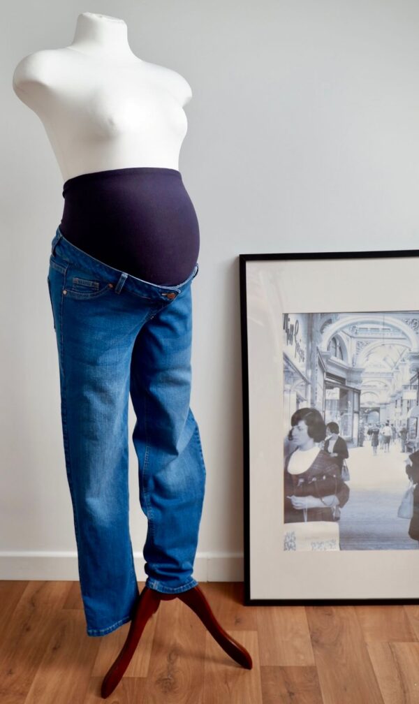 Pregnant mannequin with blue over the bump maternity jeans to the left of the imagine against a white wall. Framed picture to the right of the mannequin rests on the floor against the wall