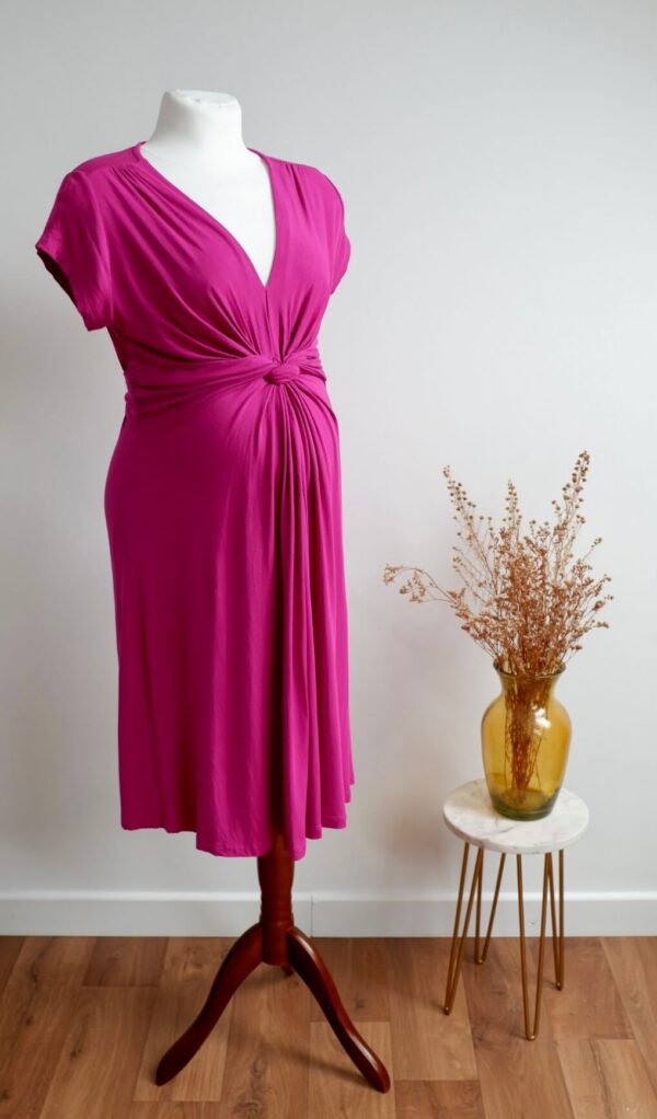 Pink maternity dress on a mannequin against a white wall with a vase with dried flowers on a table to the side