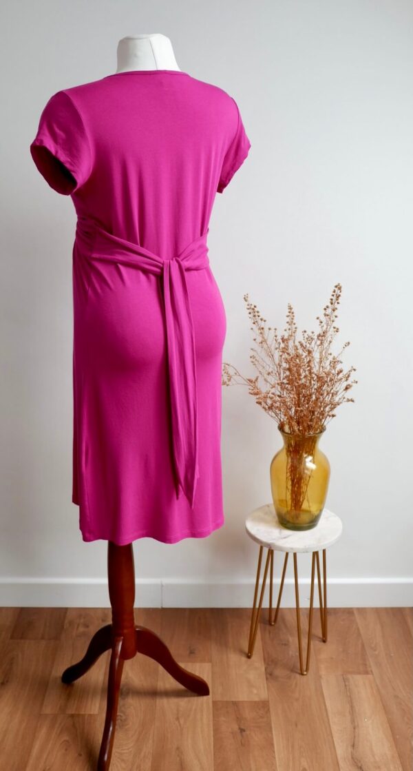 Pink maternity dress on a mannequin showing the back of the dress against a white wall with a vase with dried flowers on a table to the side