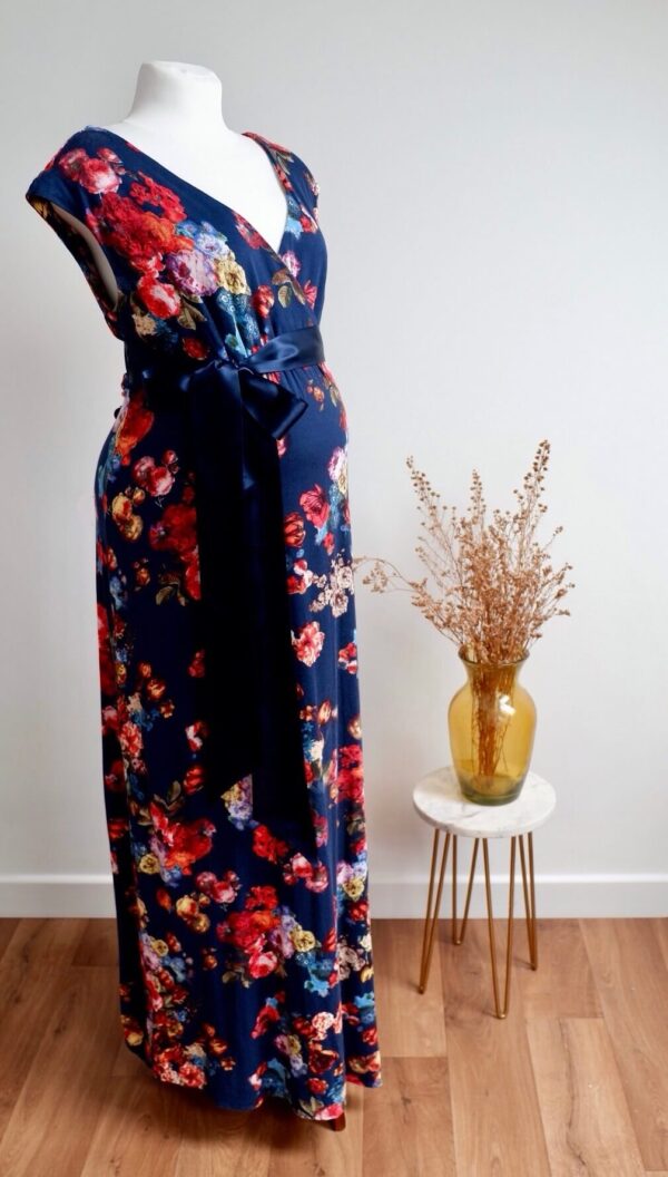 Navy floral maternity maxi dress against a white wall with a vase of dried flowers on a table to the right