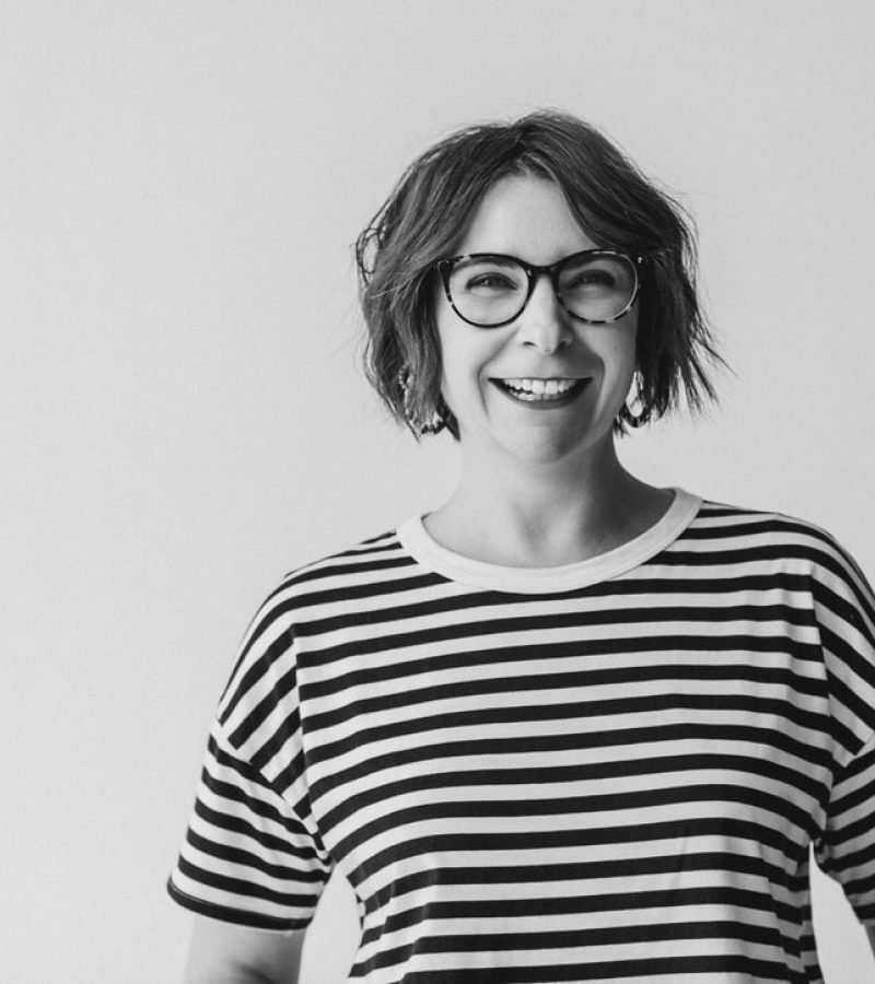 Black and white imagine of Jo Leggiero, founder of Mother Dragon standing against a wall smiling at the camera